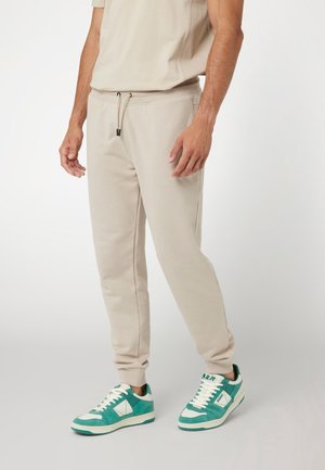 Hombre vistiendo pantalones jogger beige con cintura ajustable y zapatillas verdes y blancas, sobre un fondo claro y liso.