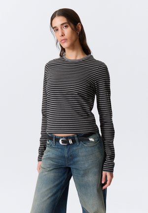 Femme aux longs cheveux foncés portant un t-shirt à manches longues rayé noir et gris et un jean bleu délavé avec une ceinture noire.