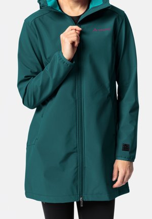 Person trägt einen dunkel-türkisfarbenen langen Mantel mit Kapuze, hält den Reißverschlussgriff nahe der Brust, Logo sichtbar auf der oberen linken Brustseite und am Ärmel.