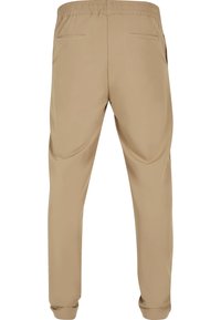 Pantalon beige décontracté avec ceinture élastique, deux poches passepoilées arrière et jambes fuselées finissant par des poignets élastiques, vue de dos.