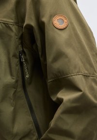 Olivengrønn jakkeerm med glidelåslomme og rundt lærmerke med teksten "Fjällräven Bergtagen".