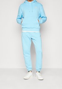 Ljusblå Nike-hoodie med känguruficka och broderad logotyp, matchad med svettbyxor i samma färg. Vita accenter och texturerad tyg. Vita sneakers.