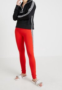 Černá mikina na zip s bílými klikatými postranními pruhy, v kombinaci s červenými legínami. Modelka má na sobě bílé sandály s barevnými pruhy.