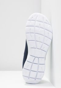 Semelle en caoutchouc blanche d'une chaussure de sport bleu marine appuyée contre un mur blanc sur une surface blanche.