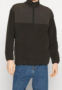 Fleece-pullover i mörkgrå, med hög krage, halvdragkedja fram och en kontrasterande överpanel med en subtil logotyp. Slät textur.