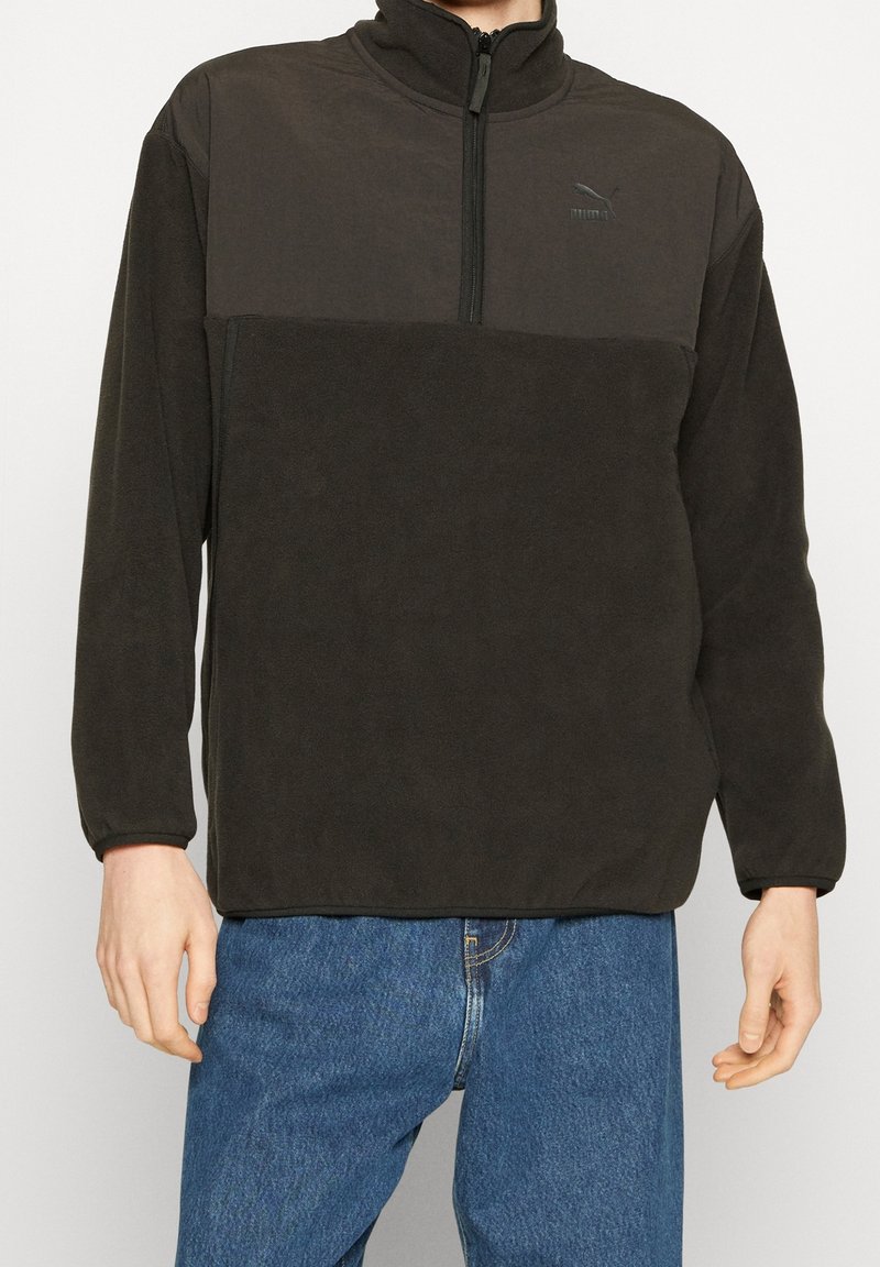 Fleece-pullover i mörkgrå, med hög krage, halvdragkedja fram och en kontrasterande överpanel med en subtil logotyp. Slät textur.