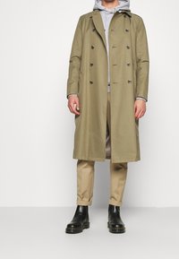 Trench coat caqui con botones de doble pecho, detallado con un cuello; llevado sobre una sudadera gris, combinado con pantalones beige y botas negras.