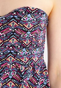 Robe sans bretelles en tissu noir avec un motif géométrique vibrant en rose, bleu, jaune et blanc. Présente un corsage ajusté et une jupe fluide.