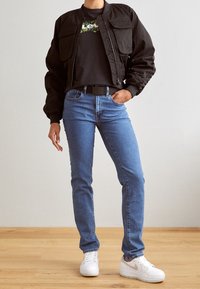 Giubbotto bomber nero, t-shirt con grafica, jeans blu e sneakers bianche. Il giubbotto ha due tasche frontali e una chiusura con cerniera.