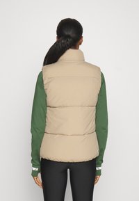 Veste sans manches matelassée beige, col montant, associée à une chemise verte à manches longues. Leggings noirs en bas.