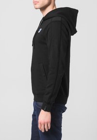 Felpa con cappuccio nera con tasca frontale, coulisse e cuciture laterali. Il tessuto sembra morbido con una texture liscia. Piccolo logo sul petto.