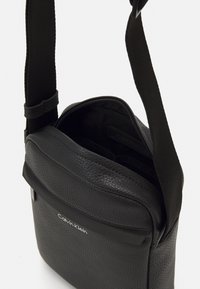 Borsa a tracolla in pelle nera con superficie texturizzata, chiusura con zip, tracolla in tessuto regolabile e logo visibile sulla parte anteriore.