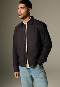Dunkelblaues, leichtes Jackett mit Reißverschluss, spitzem Kragen und Seitentaschen. Gestylt über einem beigen Hemd und hellblauen Jeans.