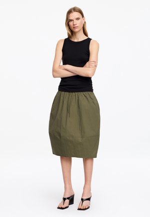 Femme debout, bras croisés, portant un haut noir sans manches, une jupe longueur genou vert olive et des sandales compensées noires sur fond blanc.