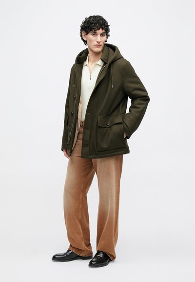 Oliivinvihreä hupullinen takki, jossa on kaksi etutaskua, beige vetoketjullinen pikeepaita ja vaaleanruskeat leveälahkeiset housut. Mallilla on mustat kengät.