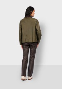 Oliwkowa skórzana kurtka z brązowym zamszowym kołnierzem, luźny krój, widoczne szwy, w połączeniu z brązowymi skórzanymi spodniami i jasnymi butami.