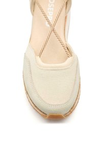 Botte en toile beige avec un bout arrondi, présentant des lacets bruns entrecroisés. La semelle texturée et les accents contrastants soulignent son design.