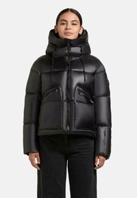 Vrouw in een glanzende zwarte pufferjack met een hoge kraag, voorrits, koordjes en ritszakken, gecombineerd met zwarte broek.