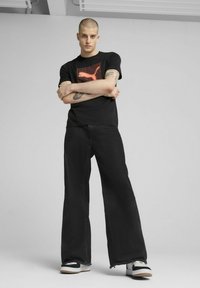 T-shirt nera in cotone con logo a griglia rossa, abbinata a jeans neri a gamba larga e sneakers bianche, nere e grigie.