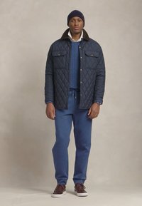 Marineblå quiltet jakke med kordfløjlskrage, blå sweater og matchende joggingbukser. Brune sneakers fuldender outfittet med et afslappet design.