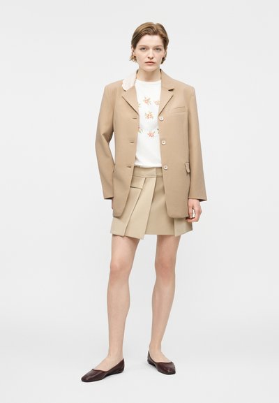 Νεαρή γυναίκα που φοράει μπεζ σακάκι, φούστα με πτυχώσεις στο ίδιο χρώμα, λευκό τοπ με λουλούδια και σκούρα καφέ επίπεδα παπούτσια, στέκεται μπροστά σε απλό φόντο.