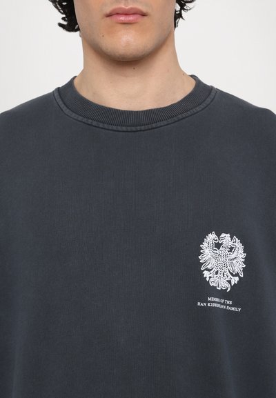 Sudadera gris oscuro con escote redondo, puños de canalé y un emblema de águila bordado en blanco con texto acompañante en el pecho.