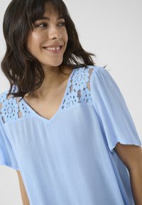 Blouse bleu clair en tissu texturé, avec un col en V et des accents de dentelle florale aux épaules. Manches courtes et fluides.