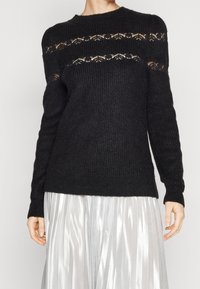 Maglione nero lavorato a maglia con girocollo, caratterizzato da dettagli a rete aperta sul petto e sulle spalle, abbinato a una gonna plissettata argentata.