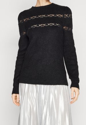 Pull en tricot noir avec un col rond, présentant des détails en maille ouverte au niveau de la poitrine et des épaules, associé à une jupe argentée plissée.