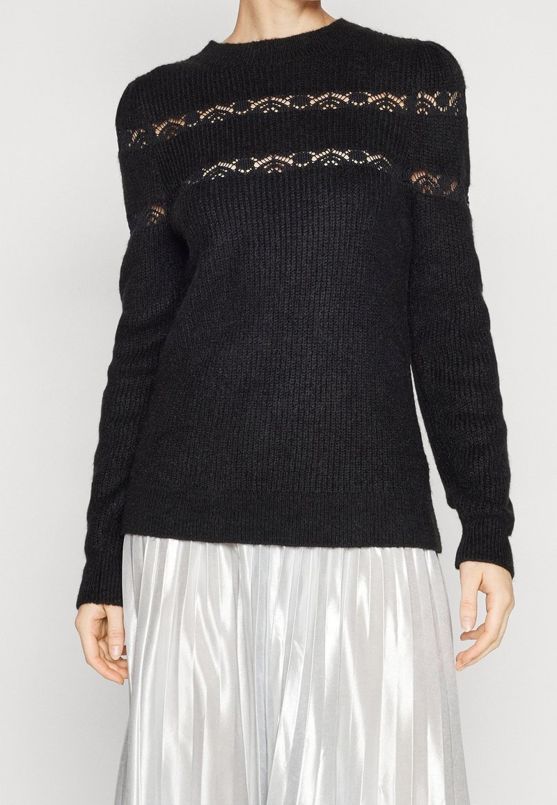 Maglione nero lavorato a maglia con girocollo, caratterizzato da dettagli a rete aperta sul petto e sulle spalle, abbinato a una gonna plissettata argentata.