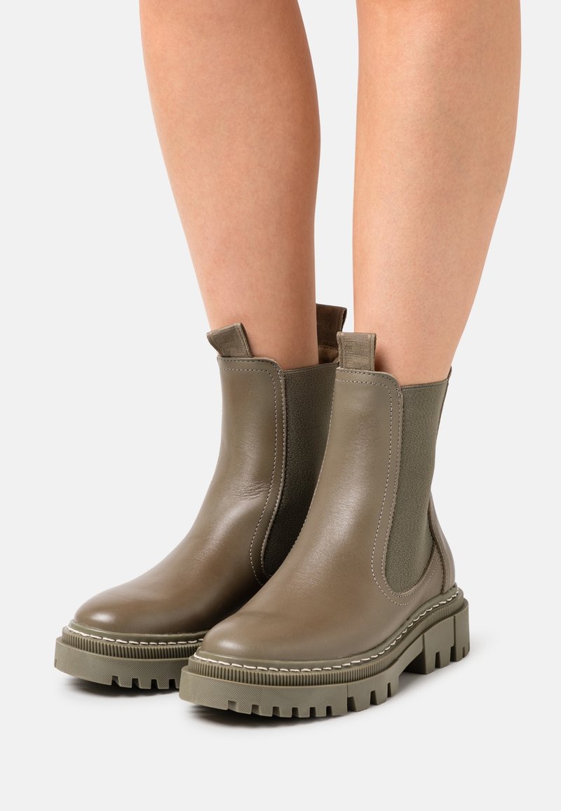 Bottes chevilles en cuir vert olive avec panneaux élastiques sur les côtés, languettes de traction et semelles épaisses à crampons portées sur jambes nues.