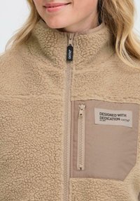 Gros plan sur une veste en polaire beige avec fermeture éclair et poche cousue étiquetée "CONÇUE AVEC DÉVOTION oxmo" portée par une personne.