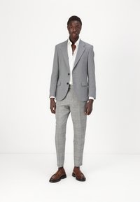 Sacou gri ajustat cu rever în formă de notch, cămașă albă și pantaloni tipici cu carouri gri deschis. Pantofi loafers din piele maro completează ținuta.