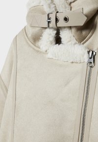 Béžová semišová bunda s měkkým bílým fleecovým límcem, vybavená kovovým zipem a ozdobným páskem na zapínání límce.