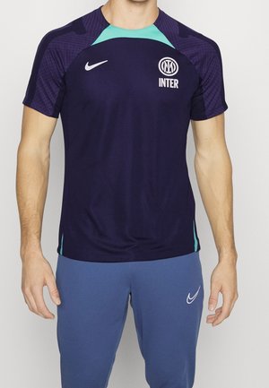 Mann trägt navyblaues und türkisfarbenes Inter Mailand Fußball-Trainingsshirt und blaue Nike-Hose, steht vor einfarbigem Hintergrund.