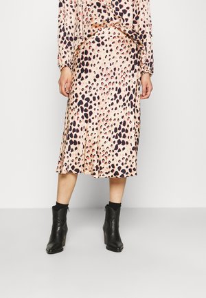 Kvinde iført en midinederdel med beige og sorte pletter, matchende bluse og sorte ankelstøvler, stående foran en ensartet hvid baggrund.