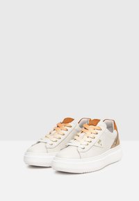 Sneaker bianche in pelle con dettagli color caramello, pannelli laterali testurizzati, suole spesse bianche e lacci arancioni fissati da occhielli dorati.