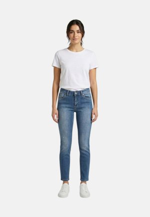 Donna in piedi su sfondo bianco, indossa una maglietta bianca semplice, jeans blu aderenti e scarpe da ginnastica bianche, rivolta in avanti con postura rilassata.