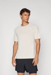 Jasno beżowy t-shirt z krótkim rękawem, okrągłym dekoltem i rękawami raglanowymi. Posiada minimalne logo w jaśniejszym odcieniu. Miękka struktura materiału.