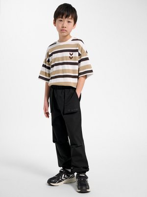 Chłopiec stoi z rękami w kieszeniach, ubrany w beżowo-brązowy koszulkę w paski, czarne spodnie i czarne sportowe buty na białym tle.