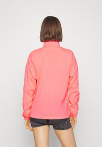 Osoba stojąca tyłem, ubrana w jaskraworóżową kurtkę z długim rękawem, z małym białym logo z tyłu oraz czarnymi sportowymi szortami w wzory.