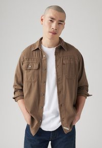 Camisa de algodón marrón con frente abotonado, dos bolsillos en el pecho y mangas remangadas, combinada con una camiseta blanca y jeans oscuros.