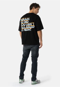 Černé tričko s krátkým rukávem se multicolor textem na zádech, který zní "SNIJTE VELKÉ, NEMŮŽETE VŠICHNI SPI T POŘÁD." Nosí se s tmavými džíny.