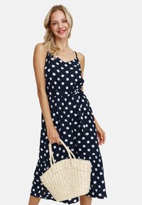 Granatowa sukienka w kropki z dekoltem w serek, regulowanymi ramiączkami i związanym w pasie. Posiada zwiewną spódnicę oraz tkany beżowy torebkę.