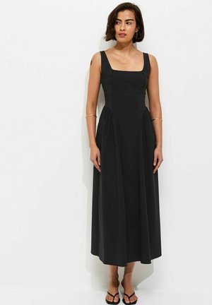 Femme debout sur fond blanc portant une robe midi noire sans manches avec un décolleté carré et des sandales noires.