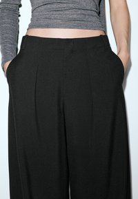 Czarne spodnie z szerokimi nogawkami o gładkiej fakturze, z przodu z zakładkami i bocznymi kieszeniami, zestawione z dopasowanym szarym długim rękawem.