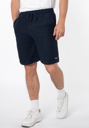Man draagt marineblauwe corduroy shorts met elastische tailleband en trekkoord, witte sokken en witte sneakers, staand tegen een witte achtergrond.