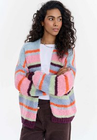 Žena s kudrnatými vlasy, oblečená v barevném pruhovaném pleteném kardiganu přes bílou košili a tmavé kalhoty, se založenýma rukama, dívající se stranou.