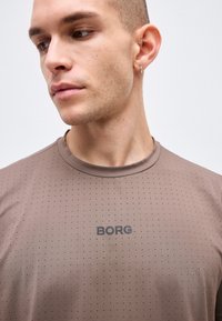 Νέος άνδρας με κοντά μαλλιά κοιτάζει αριστερά, φοράει μικρούς κρίκους και καρφωτά σκουλαρίκια και ένα καφέ πουκάμισο με τρύπες με το λογότυπο "BORG".