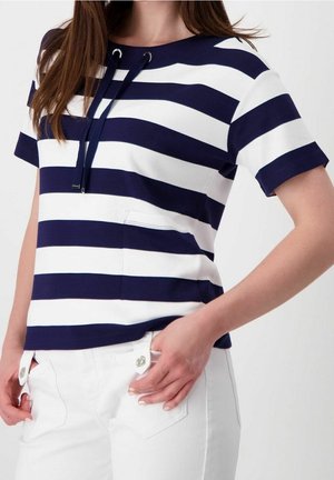 Kobieta ubrana w granatowo-biały top z krótkim rękawem w poziome paski oraz białe spodnie, ręka spoczywa na biodrze.